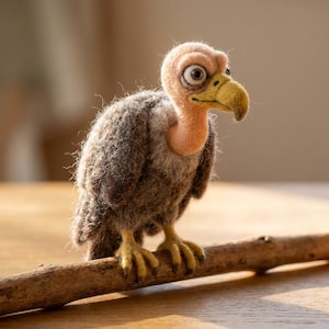 May include: A needle-felted vulture figurine with a textured dark brown and gray wool body, a smooth peach-colored head and neck, and a yellow beak. The bird has large, wide eyes and yellow feet, and it is perched on a thin, horizontal wooden branch.