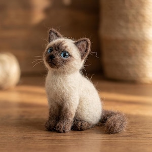 Op de afbeelding: Een naaldgevilte Siamees kittenfiguur met een crèmekleurig lichaam, donkerbruine oren, gezicht en pootjes, en helderblauwe ogen. De kleine kat zit rechtop met de staart naar voren gekruld en heeft een pluizig, getextureerd oppervlak van wol.