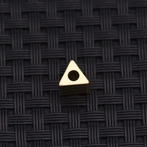 Könnte beinhalten: Eine kleine, goldfarbene Metallperle in Dreiecksform mit einer glatten, reflektierenden Oberfläche. Die Perle verfügt über ein einzelnes kreisförmiges Loch in der Mitte der dreieckigen Fläche, das zum Auffädeln auf eine Schnur oder einen Draht dient.