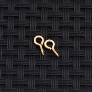 May include: Two small gold-colored metal screw eyes, each featuring a circular loop at the top and a threaded shank at the bottom, designed for fastening or hanging items. The hardware has a smooth, polished finish and a distinct spiral thread.