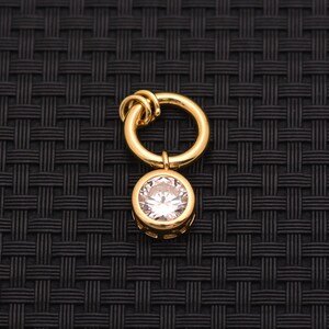 May include: A gold-toned metal charm featuring a circular ring at the top and a single round, clear faceted gemstone set in a gold-toned bezel at the bottom. The charm has a polished finish and a simple, clean design for jewelry attachment.