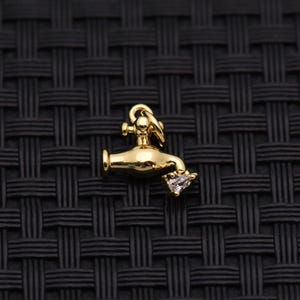 May include: A gold-toned metal charm shaped like a water faucet with a small, clear, pear-shaped gemstone set at the spout. The charm includes a circular jump ring at the top for attachment, featuring a smooth, polished finish throughout the piece.