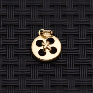 May include: A small, circular, gold-toned metal charm shaped like a four-hole button. The center features a raised, crossed-thread design connecting the four circular cutouts. A small, integrated jump ring is attached to the top for hanging.