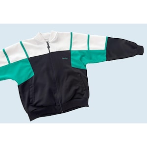 May include: A color-blocked zip-up jacket in white, black, and teal. The jacket has a stand-up collar, a full-length zipper, and a small logo on the chest. The sleeves are long and the jacket has a relaxed fit.