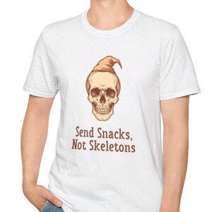Peut inclure: Un t-shirt blanc à manches courtes orné d'un graphique représentant un crâne humain portant un chapeau pointu beige. Sous le crâne, le texte "Send Snacks, Not Skeletons" est imprimé dans une police marron à empattements, centré sur la poitrine.