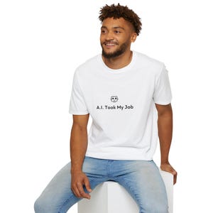 Peut inclure: Un t-shirt blanc à manches courtes arborant une petite icône de robot gris centrée sur la poitrine au-dessus du texte noir A.I. Took My Job. Le vêtement est associé à un jean en denim bleu clair, créant une apparence décontractée et simple.
