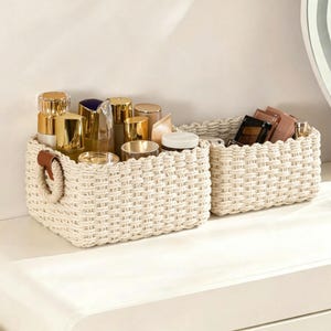 May include: Two rectangular woven baskets made of off-white rope with a thick, textured weave. The larger basket features a brown leather tab detail on the side handle. Both baskets are filled with various cosmetic containers and makeup palettes.
