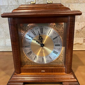 May include: A wooden mantel clock with a dark brown finish, featuring a gold-toned circular face with black Roman numerals and ornate corner filigree. The clock has a brass-colored handle on top and the text Howard Miller printed on the dial.