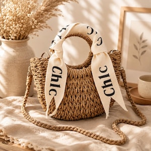May include: A woven straw handbag with a semi-circular shape, featuring two round handles wrapped in a cream-colored fabric ribbon printed with the black text "Chic". The bag includes a matching braided straw shoulder strap and a textured finish.