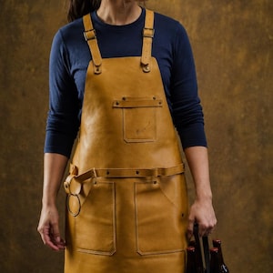 May include: A tan leather apron featuring adjustable shoulder straps with metal buckles, a chest pocket with a signature, two large waist pockets, and a side loop for tools. The apron is worn over a navy blue long-sleeve shirt.
