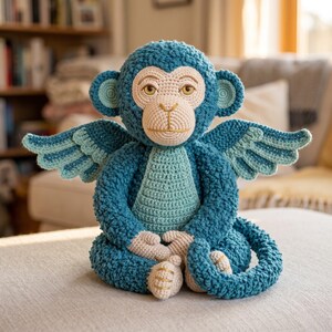 May include: A crocheted stuffed monkey toy featuring dark blue textured yarn, a light blue chest panel, and light beige face, hands, and feet. The monkey has large ears with light blue centers, a long curled tail, and two large wings with light blue accents.