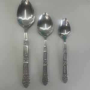 May include: Three silver-toned metal spoons of varying sizes arranged vertically. Each spoon features a polished oval bowl and a handle decorated with a repeating geometric pattern of rectangles and dots, ending in a detailed floral motif.