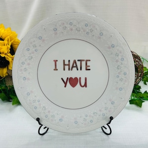 May include: A white ceramic plate featuring a delicate, embossed floral lace pattern along the rim. The center of the plate displays the text "I HATE YOU" in dark, bold lettering, with a red heart replacing the letter "O" in "you".