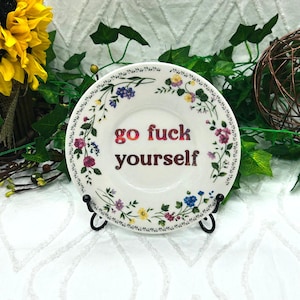 May include: A white ceramic plate featuring a floral border with small pink, blue, and yellow flowers and green leaves. The center of the plate displays the text "go fuck yourself" in a shimmering, iridescent red and dark purple font.