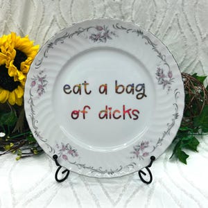 May include: A white ceramic plate with a scalloped edge and a floral border featuring pink flowers and gray leaves. The center of the plate displays the text 'eat a bag of dicks' in a multicolored, glittery, rainbow-toned font.