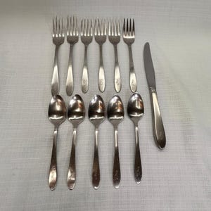 May include: A set of stainless steel flatware consisting of six forks, five spoons, and one dinner knife. Each piece features a smooth, polished finish and a small, engraved cursive letter f on the handle, arranged in two neat rows on a white surface.