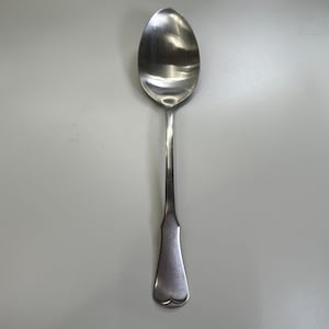 May include: A silver-colored metal spoon with a smooth, polished finish. The utensil features an oval-shaped bowl and a long, slender handle that widens slightly toward the base, which has a subtle, rounded, heart-like indentation at the tip.