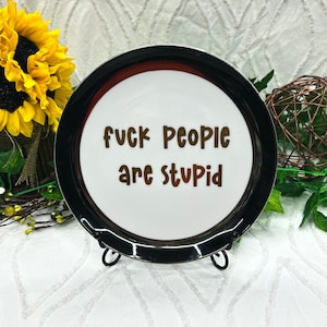 May include: A round ceramic plate with a white center and a wide black rim. The text "fuck people are stupid" is printed in brown, sans-serif letters across the center of the plate. The plate is displayed on a black metal stand.