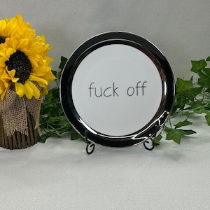 May include: A round white ceramic plate with a black rim and the text "fuck off" printed in a simple black sans-serif font. The plate is displayed on a black metal stand, positioned next to a bouquet of yellow sunflowers in a burlap wrap.