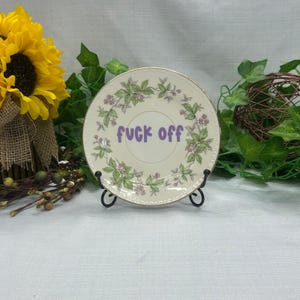 May include: A round ceramic plate with a gold-trimmed edge, featuring a decorative floral wreath pattern of green leaves and small pink flowers. The center of the plate displays the text "fuck off" in a bold, purple, sans-serif font.