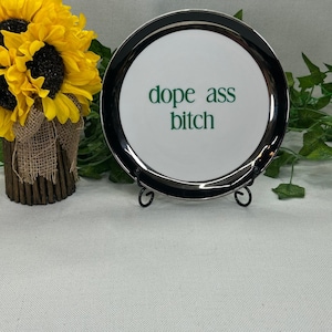 May include: A round white ceramic plate with a black rim, featuring the green text "dope ass bitch" printed in the center. The plate is displayed on a black metal stand, positioned next to a bouquet of yellow sunflowers in a burlap-wrapped vase.