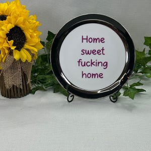 May include: A round white ceramic plate with a thick black rim, displayed on a black metal stand. The center of the plate features the purple text "Home sweet fucking home" printed in a simple sans-serif font, centered on the white surface.