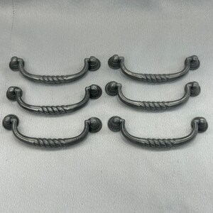 May include: A set of six dark gray metal drawer pulls arranged in two columns of three. Each pull features a curved, rope-textured handle attached to two circular mounting bases, creating a uniform appearance for furniture hardware applications.