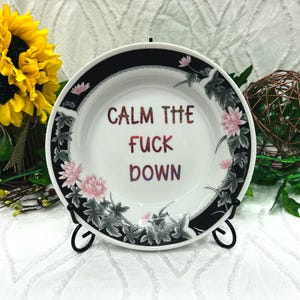 May include: A white ceramic plate with a black border featuring pink lotus flowers and grey leaves. The center of the plate displays the text "CALM THE FUCK DOWN" in a glittery, multi-colored gradient font. The plate is held by a black metal stand.