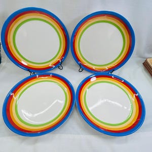 May include: A set of four round ceramic dinner plates, each featuring a white center surrounded by concentric rings in shades of bright blue, red, orange, yellow, and light green. The plates have a smooth, glossy finish and a wide, colorful rim.