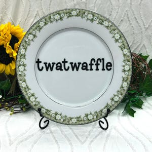 May include: A white ceramic dinner plate featuring a decorative border of small white flowers and green foliage. The center of the plate displays the word twatwaffle in bold black lowercase lettering. The plate rests on a black metal stand.
