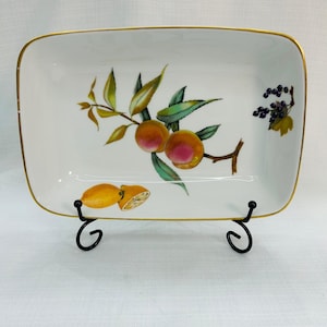 May include: A rectangular white porcelain serving dish with a gold-trimmed rim. The center features a botanical illustration of two peaches on a leafy branch, a cluster of dark purple berries, and a sliced yellow lemon at the bottom.