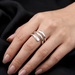 Könnte beinhalten: Ein polierter Ring in Silberoptik mit einem Bypass-Design, bei dem zwei überlappende Bänder um den Finger gewickelt sind. Das Metall hat eine glatte, reflektierende Oberfläche mit abgerundeten Enden, die eine durchgehende Spiralform bilden.
