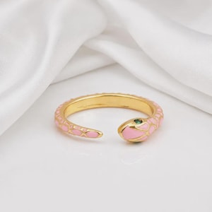 Könnte beinhalten: Ein goldfarbener, offener Ring in Schlangenform mit einer glatten, rosafarbenen Emaille-Einlage am Körper und einem kleinen, runden grünen Stein als Auge. Das Band ist flexibel, lässt sich anpassen und zeigt ein detailliertes Schuppenmuster.