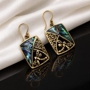 Könnte beinhalten: Ein Paar goldfarbene Ohrhänger mit rechteckigem Rahmen, einer Einlage aus irisierendem Abalone-Muschelmaterial und einem ausgeschnittenen Libellenmotiv. Die Ohrringe haben geschwungene Ohrhaken und ein detailliertes Rankenmuster.