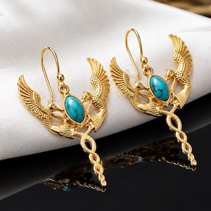 Könnte beinhalten: Ein Paar goldfarbene Ohrhänger mit einem zentralen ovalen Türkisstein, der zwischen zwei symmetrischen geflügelten Figuren sitzt. Unter dem Stein bildet das Metall ein gedrehtes, schlangenartiges Design. Jeder Ohrring hängt an einem einfachen Haken.