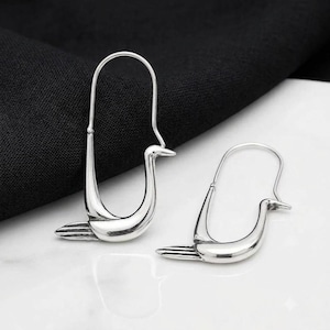 Könnte beinhalten: Ein Paar silberfarbene Ohrringe in Form stilisierter Vögel. Jeder Ohrring besteht aus einer langen, geschwungenen Drahtschlaufe, die den Körper und Hals bildet, mit einem spitzen Schnabel und einem kleinen, horizontalen Schwanzteil an der Basis.