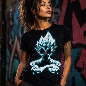 May include: A black short-sleeved t-shirt featuring a glowing light blue graphic of a spiky-haired character with a red M symbol on the forehead. The character is depicted from the chest up with arms crossed, rendered in a neon-style outline.