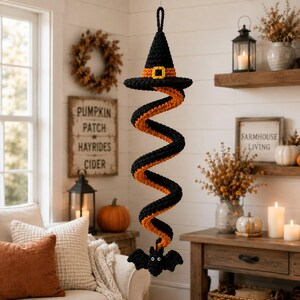 May include: A hanging crochet decoration featuring a black witch hat with an orange band and yellow buckle at the top, connected to a long, spiraling black and orange striped tail that ends with a small black bat figure with white eyes.