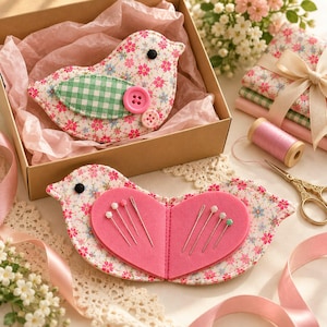 May include: Two bird-shaped fabric pincushions featuring a floral pattern in pink, blue, and green. One pincushion is inside a cardboard box with pink tissue paper, while the other is open, revealing a pink heart-shaped felt interior with needles.