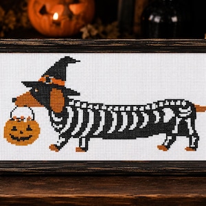 Puede incluir: Un bordado en punto de cruz enmarcado que muestra a un perro salchicha con un sombrero de bruja negro y un disfraz de esqueleto, llevando un cubo de dulces de calabaza naranja en la boca sobre un fondo de cuadrícula blanca.
