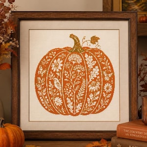 May include: A framed cross-stitch embroidery featuring a large orange pumpkin centred on a cream-coloured fabric. The pumpkin is decorated with intricate white botanical patterns, including flowers, leaves, and a small bird, with a green stem.