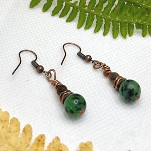 May include: A pair of drop earrings featuring round green and black speckled stone beads suspended from copper-toned wire hooks. The beads are secured with coiled copper wire accents, creating a textured look with earthy tones and metallic elements.