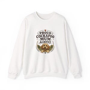 Op de afbeelding: Een wit sweatshirt met lange mouwen en een ronde hals, voorzien van een centrale afbeelding van een bruine Cockapoo-hond. De tekst PROUD COCKAPOO MUM ALWAYS staat in een cirkelvormig kader met kleine pootafdrukken, sterren en een hartje.