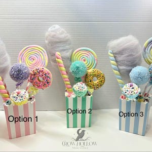 May include: Three decorative candy-themed centerpieces in striped containers labeled Option 1, Option 2, and Option 3. Each features faux cotton candy, a spiral lollipop, a donut, and a cupcake on sticks. The containers are pink, green, and blue.
