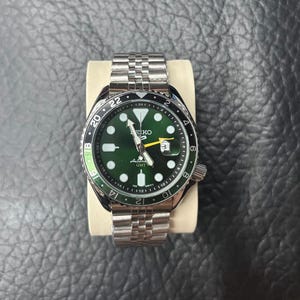 Pode incluir: Um relógio de pulso automático Seiko 5 GMT em aço inoxidável com mostrador verde escuro, marcadores de hora luminescentes, janela de data na posição das três horas, ponteiro GMT amarelo e bisel rotativo preto e verde com bracelete jubilee.