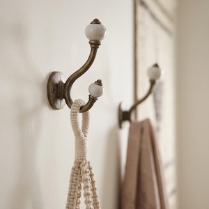 May include: A wall-mounted double hook featuring an antique brass finish and two smooth white ceramic knobs at the ends. The curved metal structure provides two points for hanging items, with a cream-coloured macrame cord looped over the lower hook.