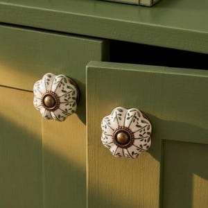 May include: Two round, fluted ceramic cabinet knobs featuring a white base with a green leaf pattern and a central bronze-toned metal accent. The knobs are attached to a solid olive green cabinet door, showcasing a scalloped edge design.