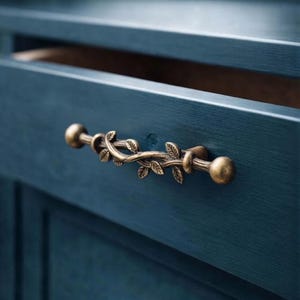 May include: A bronze-toned metal drawer pull featuring a textured vine design with small leaves, attached to a dark blue painted wooden drawer front. The handle has a rounded knob at each end and a central section shaped like a twisted branch.