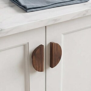 May include: Two semi-circular cabinet pulls made of dark brown wood with a visible grain pattern. The handles are mounted vertically on the front of two white cabinet doors, each featuring a flat edge and a curved outer profile for gripping.