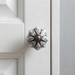 May include: A white and black ceramic cabinet knob with a faceted, star-like shape and geometric line patterns. The centre of the knob features a circular, metallic bronze-toned hardware accent, mounted on a white panelled cabinet door.